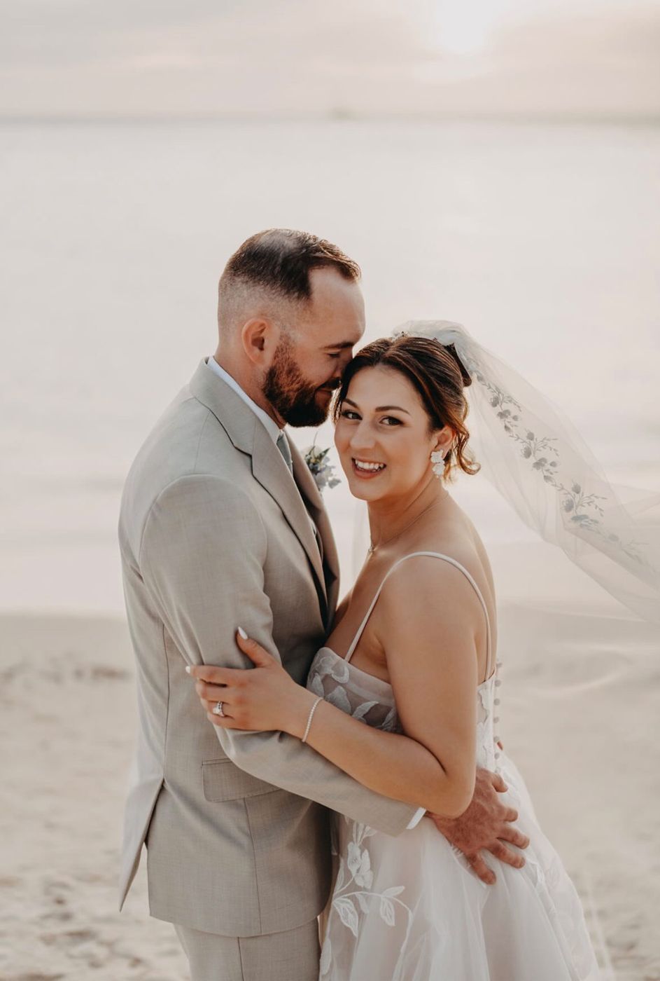 Smiling bride with a soft updo and veil in her groom's arms on the beach at sunset