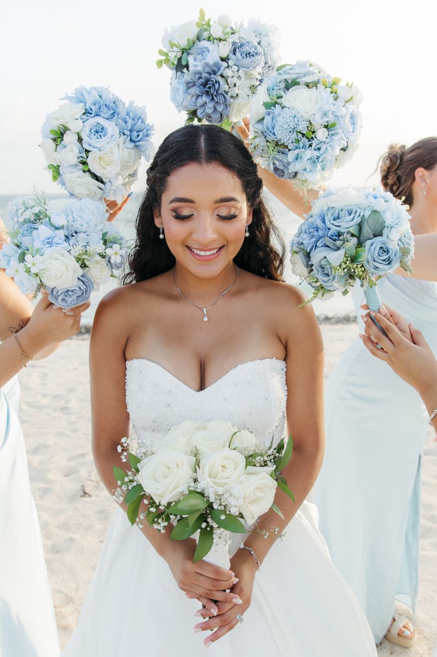 Smiling bride on the beach framed by her bridesmaids' blue bouquets