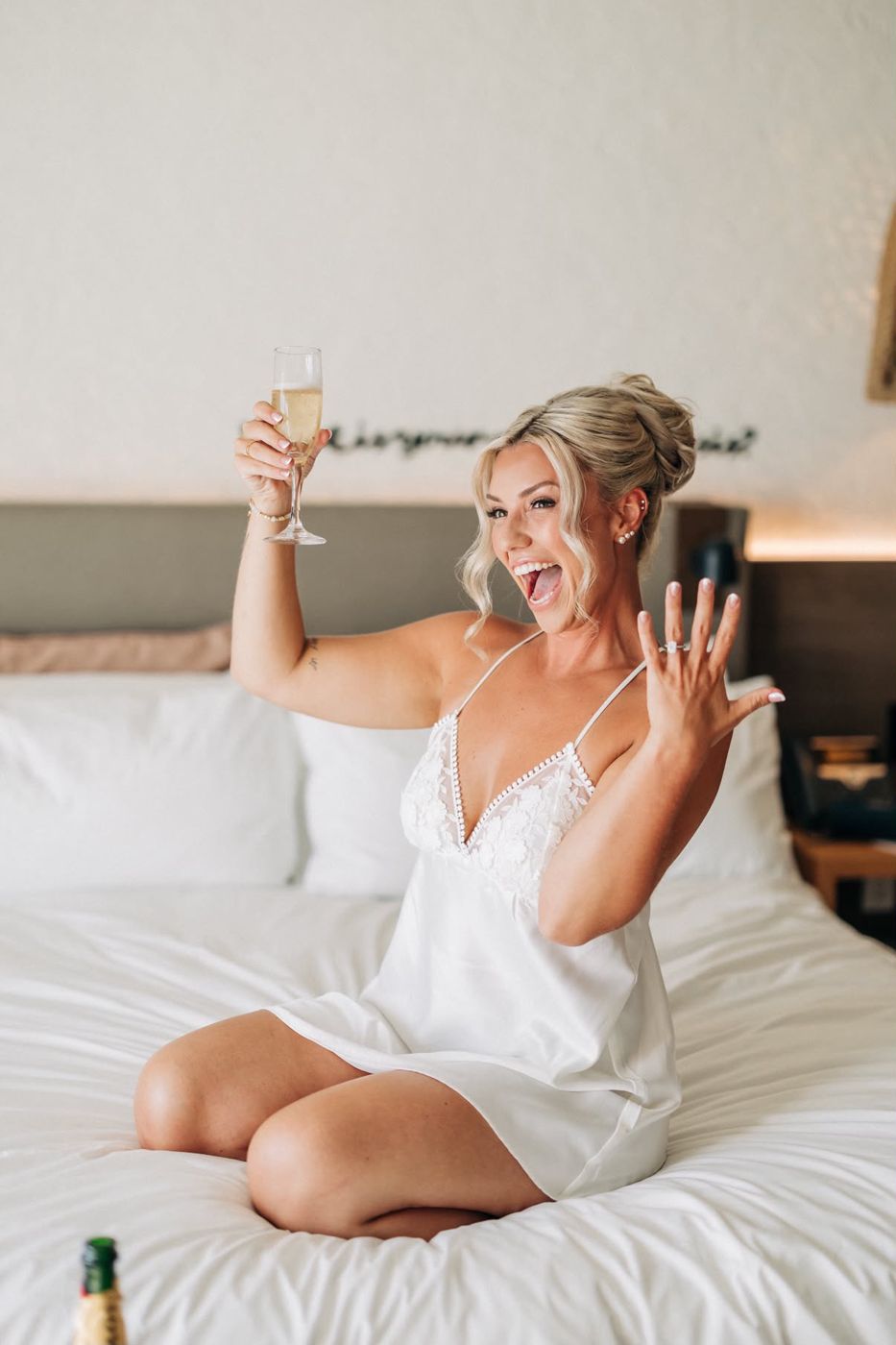 Bride on the bed raising a glass of champagne and showing her ring on her wedding morning