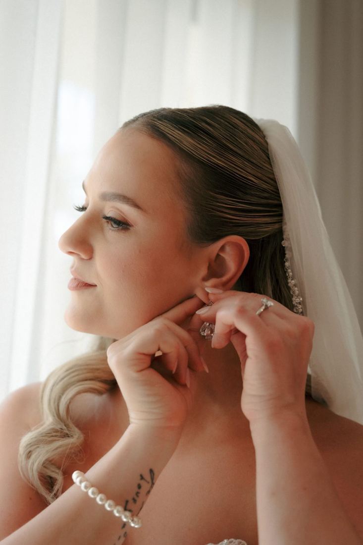 Bride putting on a drop earring in soft window light