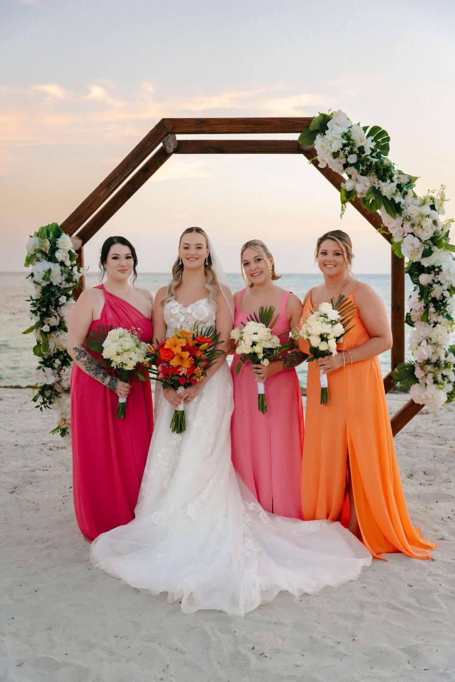 Bride with bridesmaids in pink and orange under a floral arch on the beach