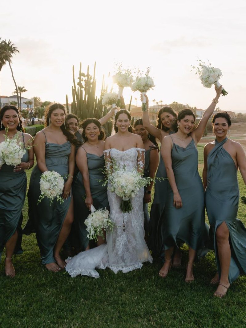 Bride and eight bridesmaids in sage satin cheering with their bouquets at sunset